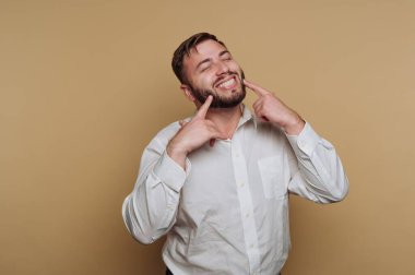 Genç, neşeli bir adamın portresi, büyük bir gülümsemeyle, yanaklarını gösteren, bej arka planda beyaz bir gömlek giyen, neşeli bir poz verir.