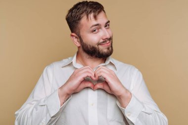 Genç bir adamın portresi bej arka planda elleri ile bir kalp şekli oluşturur.