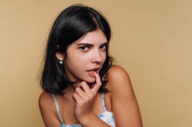 Genç bir kadının portresi, dudaklarında neşeli bir parmak ile baştan çıkarıcı bir poz veriyor, bej arka planda gizemli bir cazibe yaratıyor.