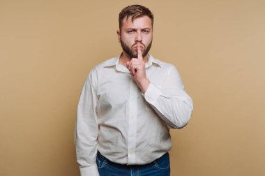 Dudaklarına parmağını dayamış genç bir adamın portresi, sessizlik işareti veriyor, bej arka planda gizem ve gizliliği somutlaştırıyor
