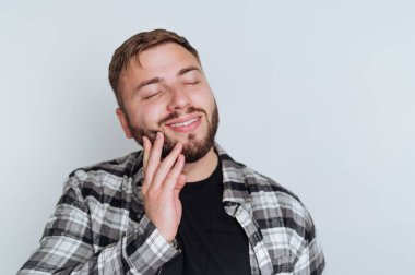 Sakallı neşeli bir gencin portresi sevinçle gülümsüyor, beyaz arka planda memnuniyetini ve memnuniyetini gösteriyor.