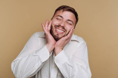 Neşeli genç bir adamın portresi, parlak bir gülümseme, bej arka planda mutluluk ve memnuniyet sergiliyor