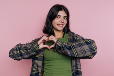 Elleriyle kalp şekli oluşturan neşeli genç bir kadının portresi pembe arka planda sevgiyi ve pozitifliği simgeliyor.