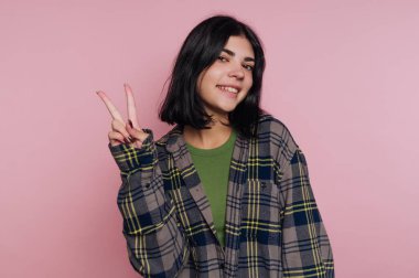 Koyu renk saçlı genç bir kadının portresi, pembe arka planda neşe ve arkadaşlığı ifade eden bir barış işareti gösteriyor.