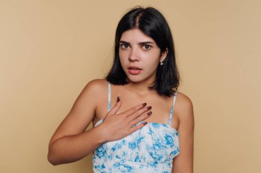 Mavi çiçekli elbiseli kızın portresi göğsünü sıkıca kavramış, bej arka planda sürpriz bir ifade sergiliyor.