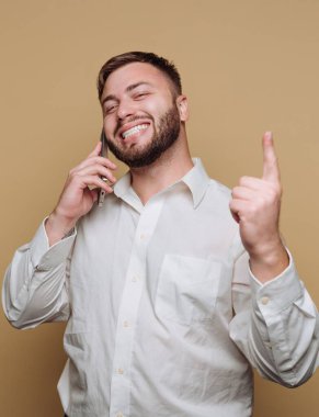 Genç bir adamın portresi, bej arka planda neşeli bir ifade sergileyen bir telefon görüşmesinde heyecan sergiliyor.