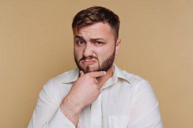 Düşünceli bir ifadeyle genç bir adamın portresi bej arka planda çenesine dokunurken pozlar veriyor.