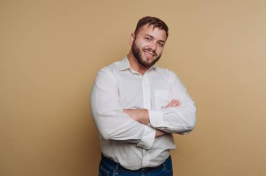 Genç gülümseyen bir adamın portresi, bej arka planda beyaz bir gömlek içinde kollarını kavuşturarak güven içinde poz veriyor.
