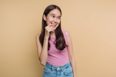 Pembe bluzlu, kot pantolonlu, bej arka planda gülümseyen genç bir kadın..