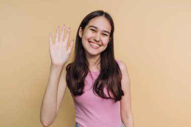 Gülümseyen genç bir kadın bej bir arka plana el sallıyor..