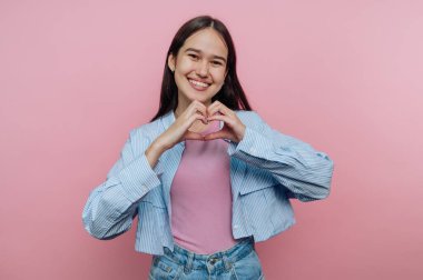 Gülümseyen genç bir kadın pembe bir arka planda elleriyle kalp şekli oluşturuyor..