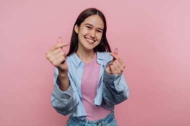 Gülümseyen genç bir kadın pembe arka planda parmaklarıyla kalp hareketleri yapıyor..