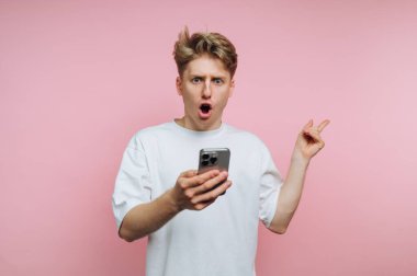 Pembe arka planda akıllı telefonu tutan şaşkın suratlı genç bir adam..