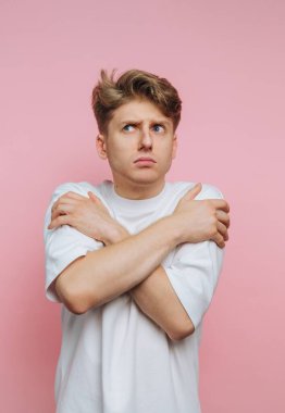 Beyaz tişörtlü genç adam pembe arka planda kollarını kavuşturuyor.