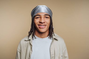 Saçları örgülü ve diş telleri bej arka planda gülümseyen genç bir adam..