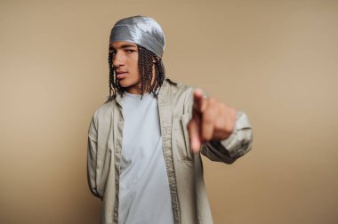 Saçı örgülü genç bir adam. Durag ve günlük giysiler giyiyor. Bej arka planda kameraya doğru bakıyor..