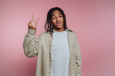 Örgülü genç adam pembe arka planda barış işareti yapıyor..