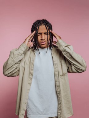 Saçları örgülü, başı pembe arka planda rahatsızlık içinde olan biri..