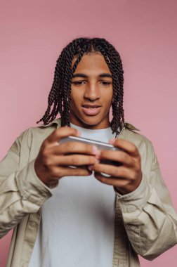 Saçları örgülü genç bir adam pembe arka planda akıllı telefon kullanıyor..