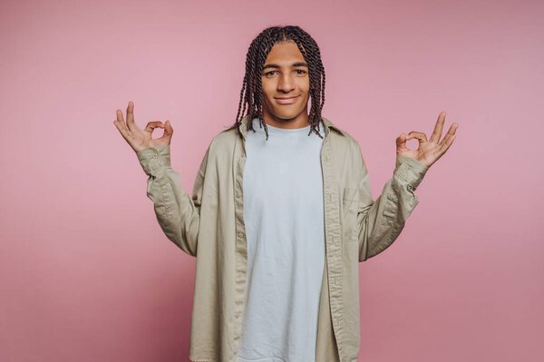 Young man with braided hair making OK signs on pink background.
