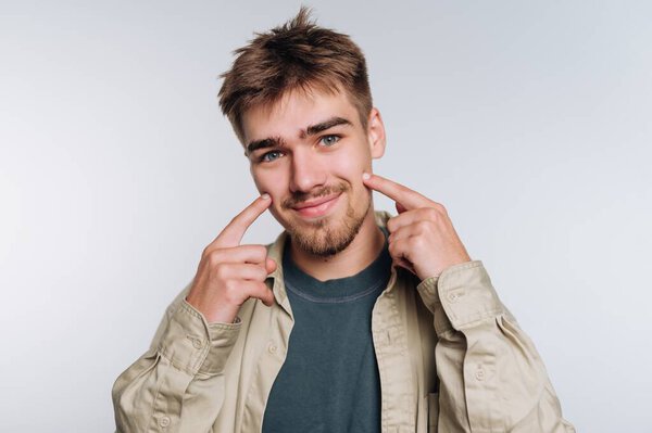 Young man smiling and pointing to his cheeks against a plain background.