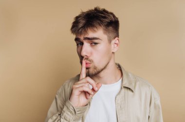 Parmakları dudaklarında genç adam sessizlik işareti veriyor, bej arka plan