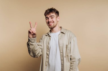 Genç adam gülümsüyor ve bej arka planda barış işareti gösteriyor.