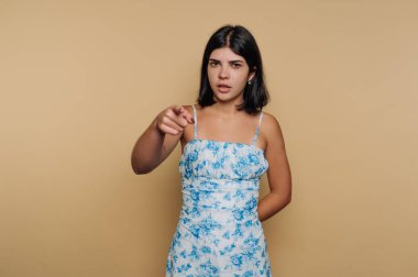 Çiçekli elbiseli genç bir kadın bej arka planda kameraya bakıyor..