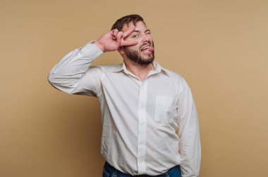 Beyaz gömlekli bir adam parmaklarıyla bir barış işareti yapıyor ve bej bir arka plana dilini uzatıyor..