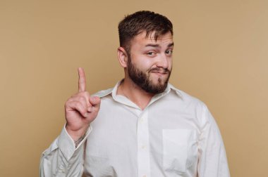 Beyaz gömlekli sakallı bir adam işaret parmağıyla bej bir arka plana doğru hareket eder..