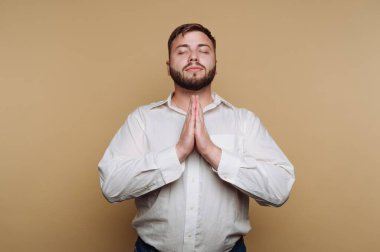 Beyaz tişörtlü, sakallı bir adam, elleri dua pozisyonunda, gözleri kapalı, bej bir arka plana karşı duruyor.