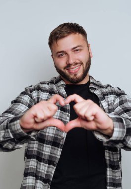 Elleriyle kalp şekli yapan gülümseyen genç bir adam, açık bir arkaplanda ekose gömlek giyiyor.