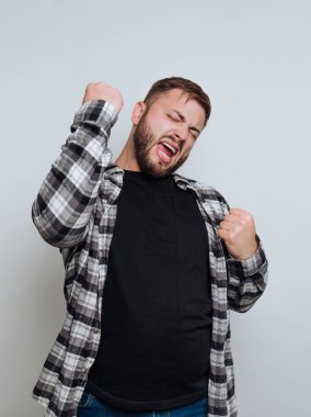 Neşeli bir ifadeyle kutlayan genç bir adam, ekose gömlek ve siyah tişört giyiyor, yumruklar heyecan içinde arka plana doğru yükseliyor.