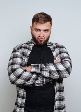 Sakallı, ekose tişörtlü ve siyah tişörtlü genç bir adam, kollarını kavuşturuyor ve düz bir arka plana karşı hoşnutsuz bir yüz ifadesi takınıyor.