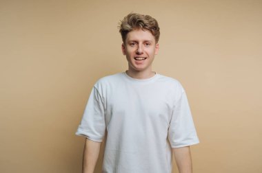 Beyaz tişörtlü genç adam bej arka planda gülümsüyor..