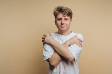 Beyaz tişörtlü genç adam bej arka planda kendine sarılıyor..