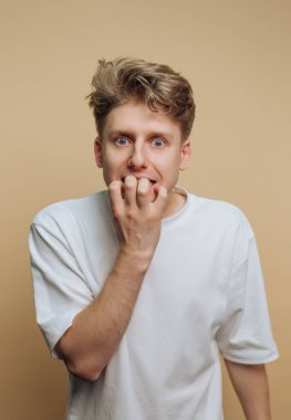 Saçları dağınık genç bir adam endişeli görünüyor, tırnaklarını yiyor, bej bir arka planı var.