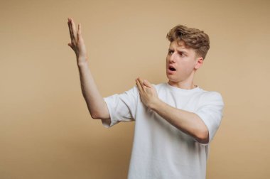 Beyaz gömlekli genç adam bej arka planda şaşkınlıkla hareket ediyor..