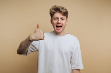 Beyaz tişörtlü genç adam bej arka plana başparmak işareti yapıyor..