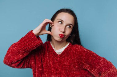 Mavi arka planı olan bir kadın içeride duruyor. Yanaklarını şişirerek ve kırmızı ruj sürerek neşeli bir ifade takınıyor. Onun kırmızı kazağı sahneye sıradan bir dokunuş katıyor..