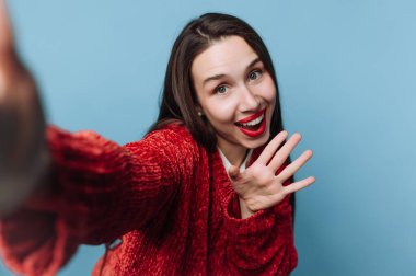Bir kadın açık mavi iç mekanda selfie çekerken gülümsüyor ve el sallıyor. Kırmızı bir kazak giyiyor ve kırmızı ruju var. Yüzünde neşe ve heyecan var..