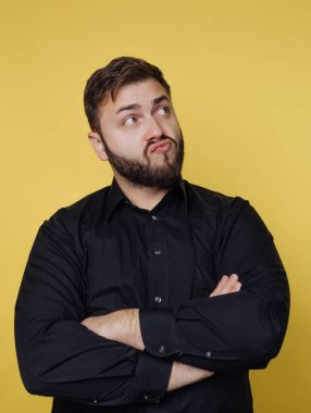 A man stands with his arms crossed and features a thoughtful expression. He has a beard and wears a black shirt. The background is yellow, and the scene is indoors.