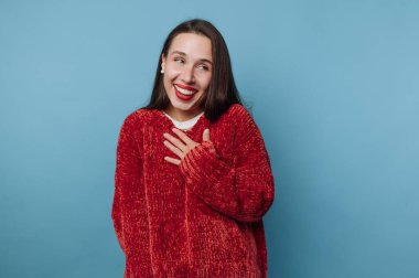 Mavi bir duvarın önünde duran genç bir kadın kırmızı bir kazak giyiyor, sıcak bir şekilde gülümsüyor. Elini göğsüne dayayıp mutlu ve meşgul görünüyor..