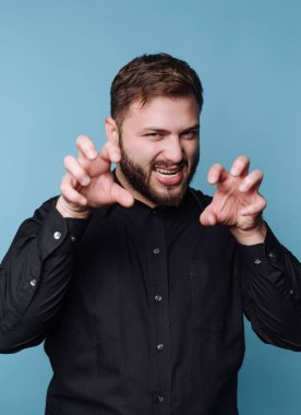 Bir adam ayağa kalkar ve elleriyle hareketli pençeler yapar. O, siyah bir gömlek giyerken gülümser. Arkaplan koyu mavi renkte..