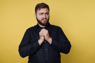 Sakallı bir adam sarı bir arka planın önünde duruyor. Ellerini birbirine kenetlerken endişeli görünüyor. Ayarlar sıradan bir atmosfer olduğunu gösteriyor..