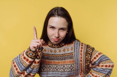 Sarı arka planda genç bir kadın duruyor. Bir parmağını kaldırır ve renkli bir kazak giyerken neşeli bir ifade takınır..