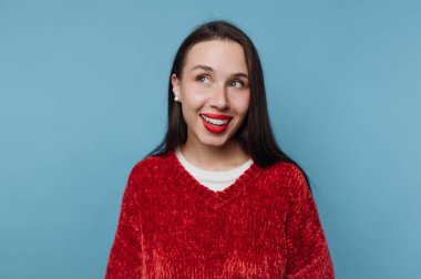 Uzun siyah saçlı bir kadın yan tarafa bakarken gülümsüyor. Kırmızı bir kazak giyiyor ve mavi bir duvara karşı parlak kırmızı bir ruju var..