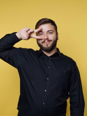 Barış işareti olarak iki parmağını kaldıran bir adam neşeli bir gülümsemeyle ayakta duruyor. Siyah bir gömlek giymiş ve parlak sarı bir duvarın önünde poz veriyor..