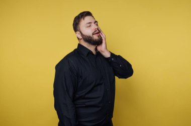 Kameraya bakan bir adam stüdyoda duruyor. Sakalı var ve siyah bir gömlek giyiyor. Sarı arka planda poz verirken yüzüne hafifçe dokunuyor..