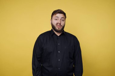 Bir adam parlak sarı bir duvara karşı sıradan bir poz verir. Sakalı var ve siyah bir gömlek giyiyor. İfadesi şakacı, komik bir anı çağrıştırıyor..